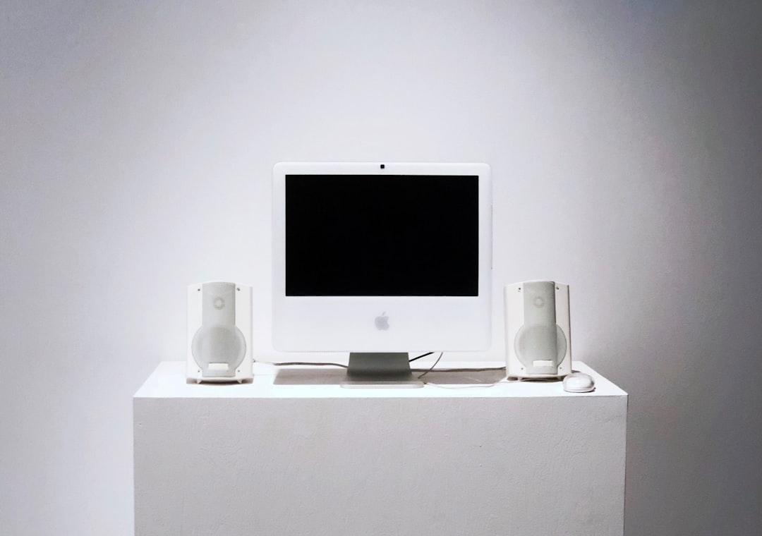 CorsiWeb.eu - Un computer desktop Apple bianco con schermo nero, due altoparlanti bianchi e un mouse esposti su un piedistallo bianco su uno sfondo bianco.